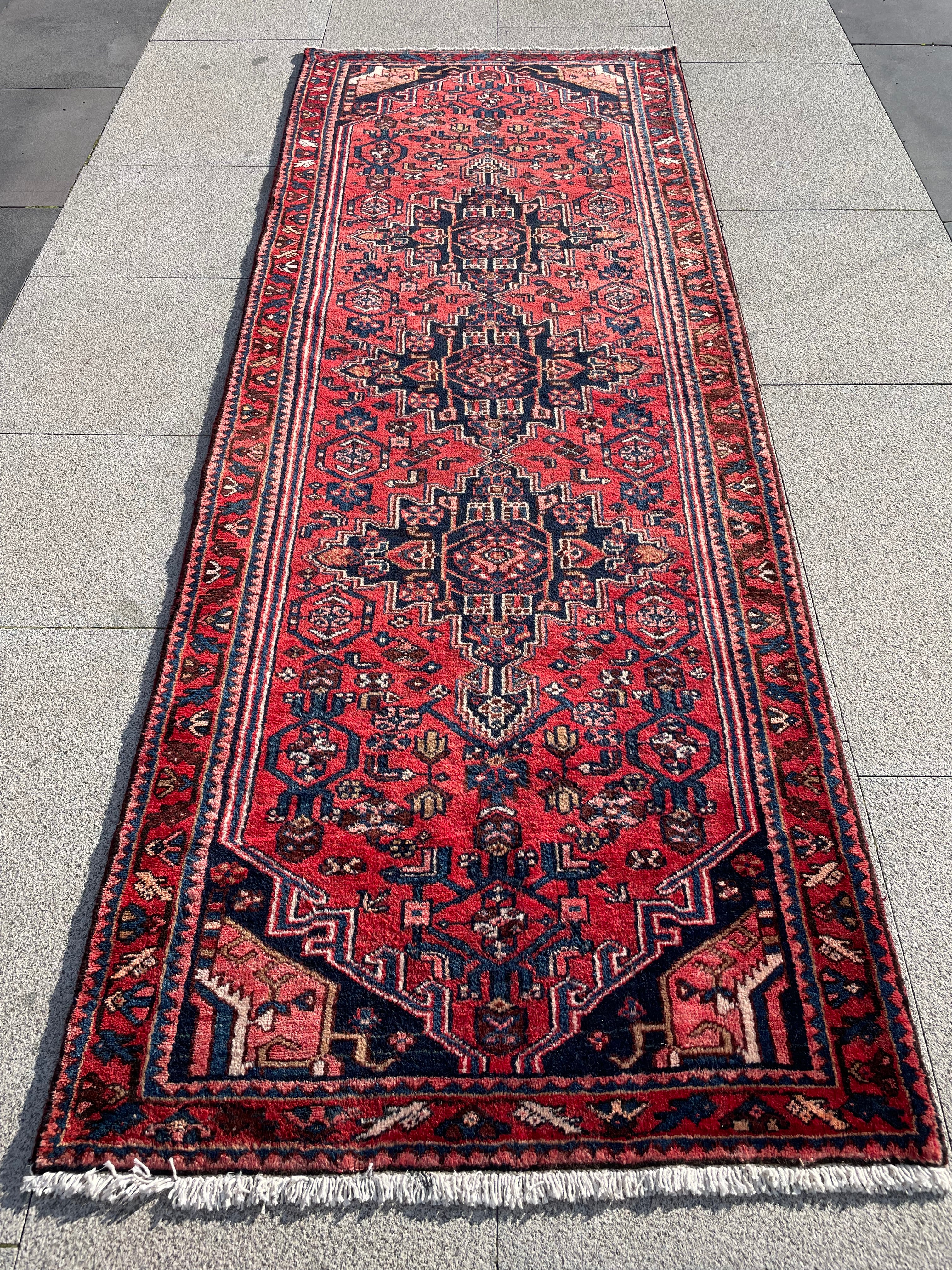 A long red Persian-style runner rug with intricate navy blue and beige geometric patterns, laid out on a light gray stone tile floor.
